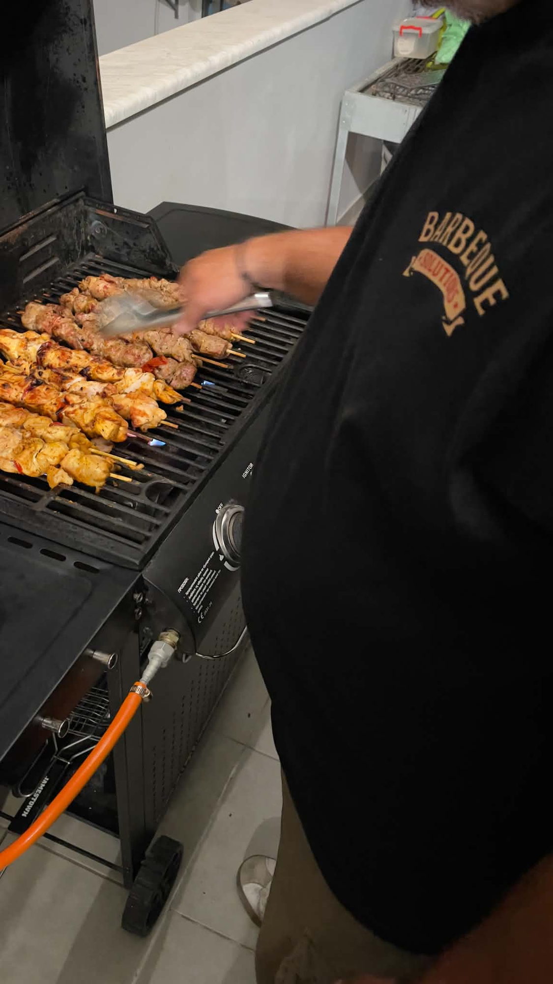 Chef in a branded shirt turning a grate full of souvlaki skewers