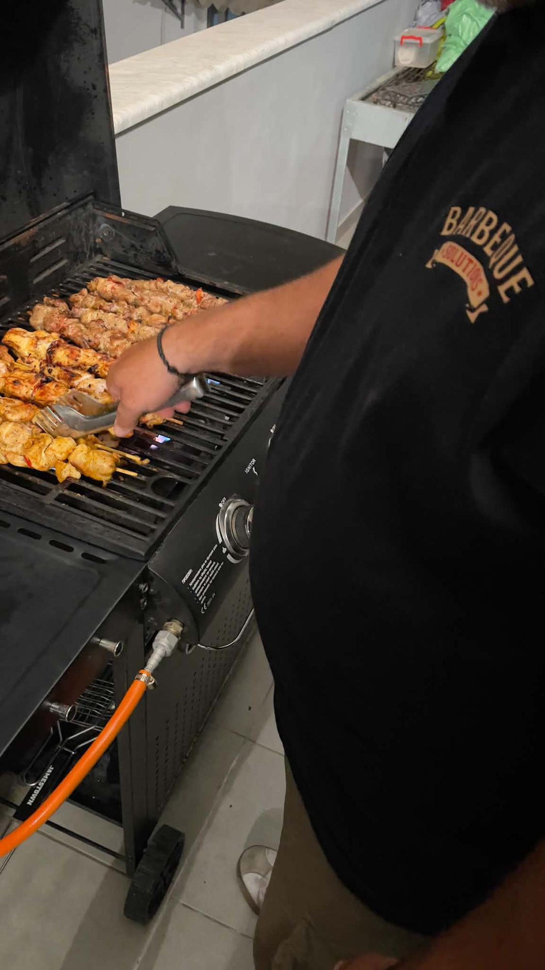 Chef turning souvlaki skewers with tongs on a gas barbecue